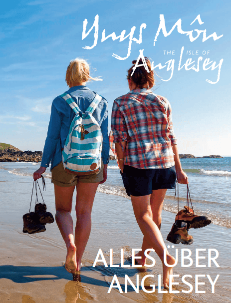 Titelblatt eines Reiseführers zu Anglesey, darauf zu sehen: 2 Frauen, die barfuß am Strand laufen, ihre Schuhe an den Schnürsenkeln in den Händen, blauer Himmel