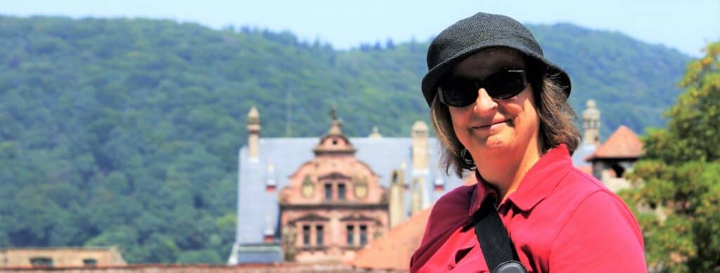 Andrea Halbritter mit Sonnenbrille, Hut und rotem Poloshirt, vor einem Schloss stehend