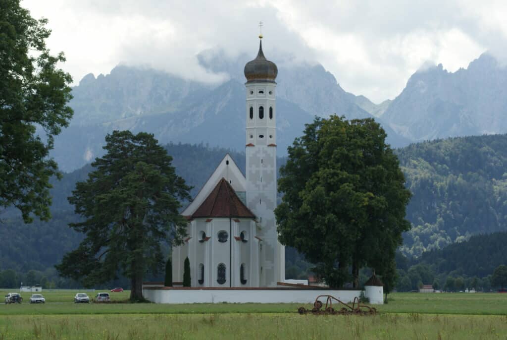 Barockkirche auf einer Wiese, davor 3 hohe Laubbäume, dahinter hohe Berge
