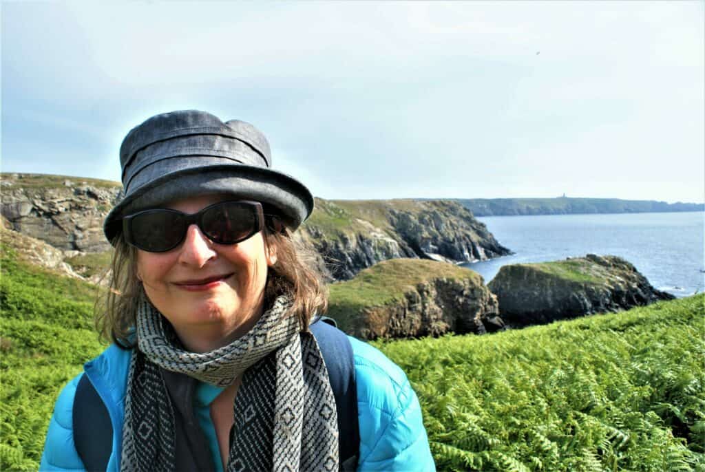 Im Vordergrund Andrea Halbritter, dahinter grüne Landspitzen und Meer, Andrea trägt eine Sonnenbrille, hat schulterlanges blondes Haar, einen Hut, eine blaue Jacke und einen Schal und lächelt in die Kamera