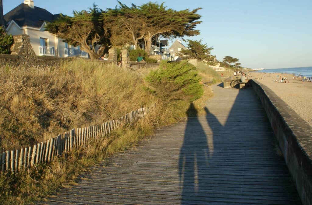 Links bewachsene Düne und Häuser, in der Mitte Weg mit Schatten von Personen, rechts Sandstrand und Meer