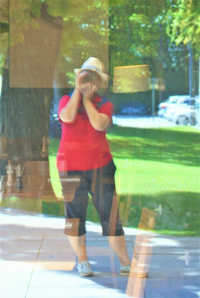 Spiegelbild von Andrea Halbritter mit Kamera in einem großen Fenster, Andrea trägt eine schwarze Hose, ein rotes Poloshirt und einen Hut.