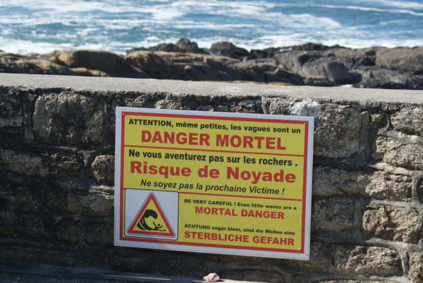 Schild auf einer Mauer vor dem Meer mit folgendem Text: Achtung, sogar klein, sind die Wellen eine sterbliche Gefahr