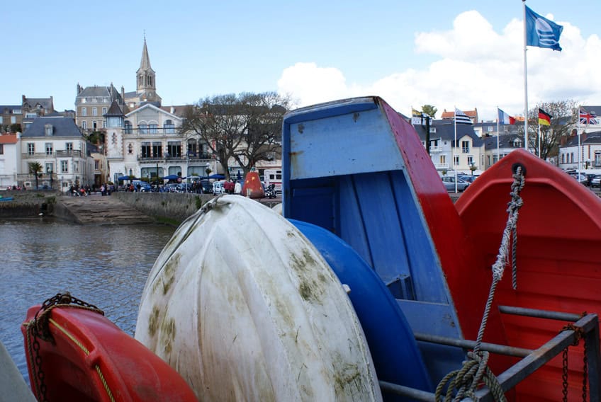Au premier blanc de petits bateaux, couleurs : rouge, blanc, bleu, derrière les bateaux de l'eau, des maisons, une église et différents drapeaux