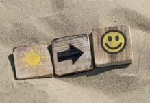 Drei Holzplättchen im Sand: auf dem 1. Plättchen eine Sonne, auf dem 2. ein Pfeil nach rechts, auf dem 3. ein zufriedener Smiley