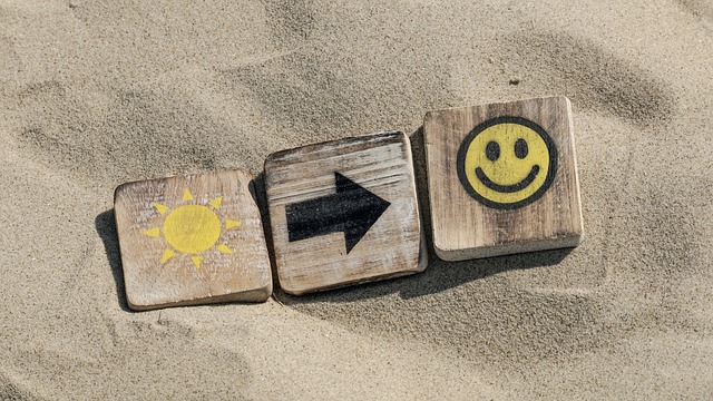 Drei Holzplättchen im Sand: auf dem 1. Plättchen eine Sonne, auf dem 2. ein Pfeil nach rechts, auf dem 3. ein zufriedener Smiley