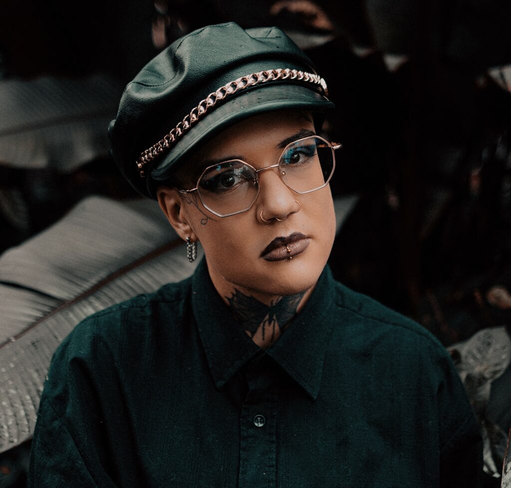 Person mit Brille, schwarzem Hemd, scharzer Kappe und Piercings in Mund und Nase sowei Schmetterlingstatoo auf dem Hals