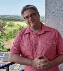 Adam Yeomans in rotem Hemd auf einem Balkon vor einer Landschaft mit Feldern und Bäumen