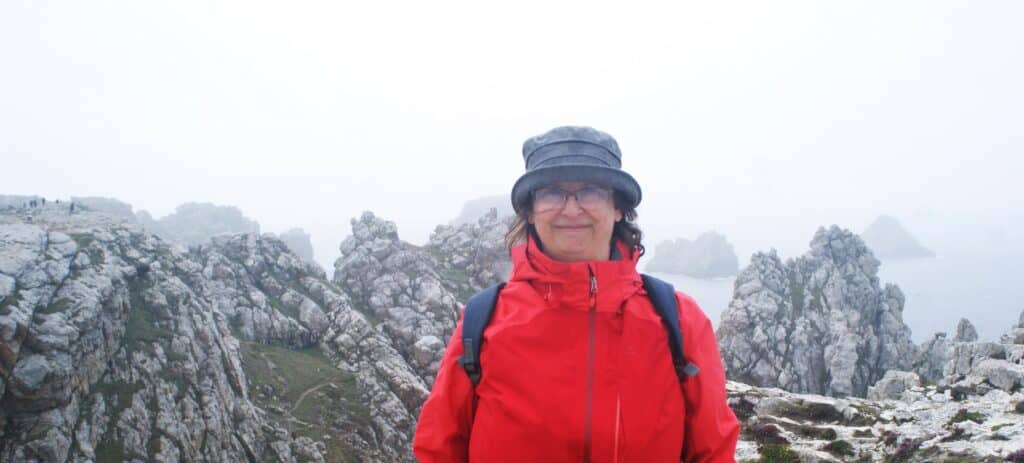 Frau mit roter Regenjacke und Regenhut vor zerklüfteter Küstenlanschaft mit Nebel und Nieselregen