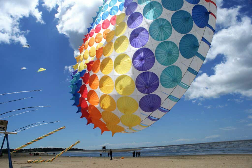 Unten im Bild ein schmaler Streifen Sandstrand mit den kleinen Silhouetten von Personen, darüber Meer und eine kleine Fischerhütte mit Senknetz, darüber wiederum viel blauer Himmel mit wenigen weißen Wolken und ein bunter runter Winddrache