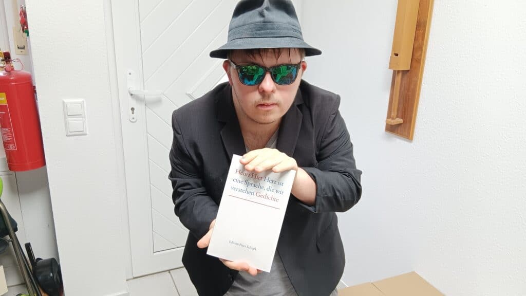 Henri mit Sakko, Sonnenbrille und Hut, in den Händen ein Buch