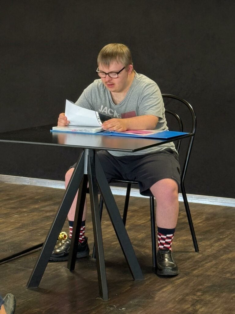 Henri sitzt während einer Autorenlesung an einem Tisch und hat einen Ordner vor sich liegen.