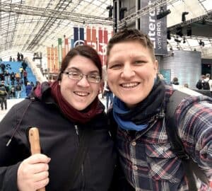 Zwei Frauen auf einer Messe; eine mit zusammengebundenen dunklen Haaren, dicker Brille und Blindenstock und eine mit kurzen Haaren