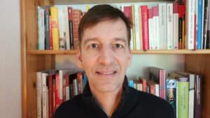 Mann mit kurzen braunen Haaren und grünen Augen vor einem Bücherregal
