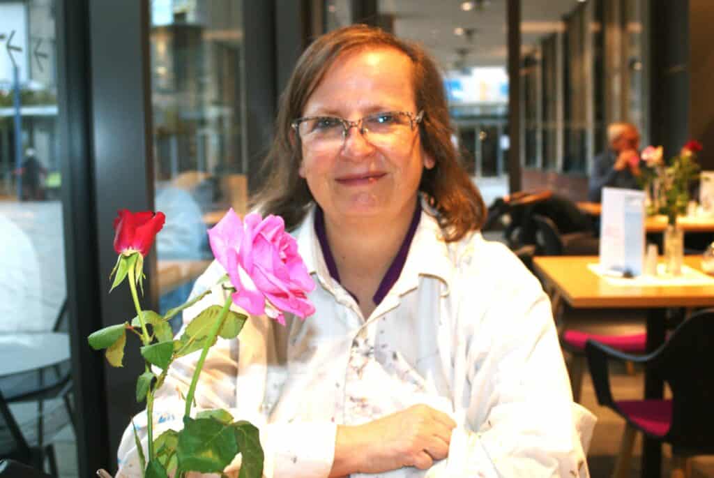 Frau mit schulterlangen dunkelblonden Haaren und Brille in einem Café. Die Frau trägt einen Malerkittel, für ihr steht eine Vase mit 2 Rosen.