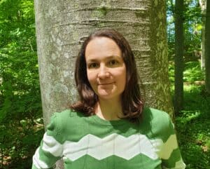 Frau mit schulerlangen braunen Haaren, dunkelbraunen Augen und weiß-grünem Pullover. Die Frau steht vor einem Baum in einem Laubwald.