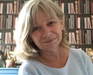 Ältere Frau mit schulterlangen hellblonden Haaren und dunkelblauen Augen. Sie trägt einen mintfarbenen Pullover und steht vor einer Bücherwand.