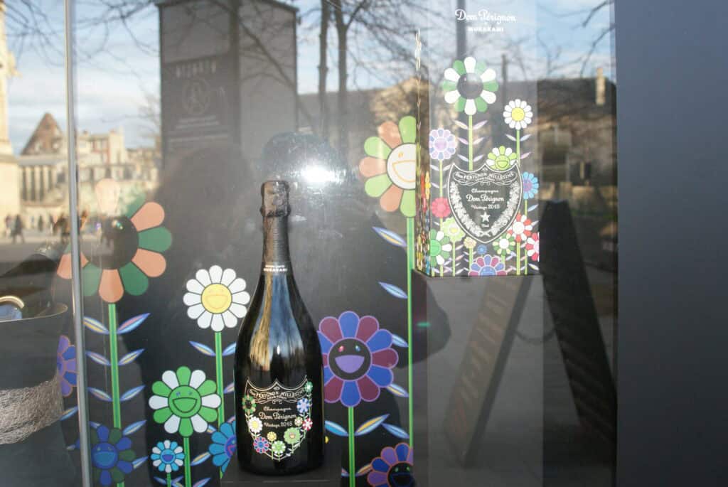 Champagnerflasche und Champagnerverpackung in einem Schaufenster mit gemalten Blumen als Dekor, im Fenster spiegeln sich historische Gebäude und die Übersetzerin