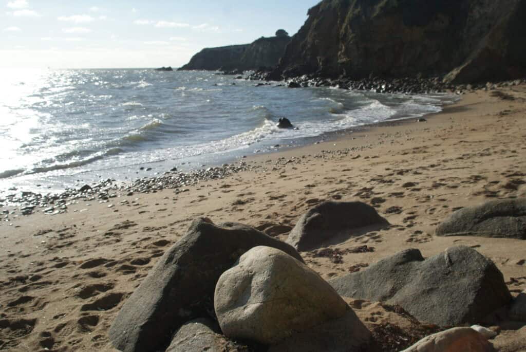 Kleine Bucht mit schmalem Sandstrand und großen Steinen, im Hintergrund hellgraues Meer mit Sonne