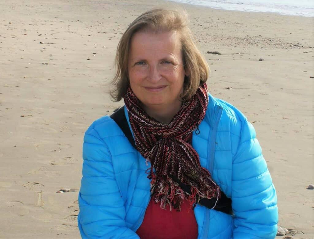 Frau mit kinnlangen blonden Haaren, blauen Augen, blauem Anorak, rotem Pullover und Schal am Strand