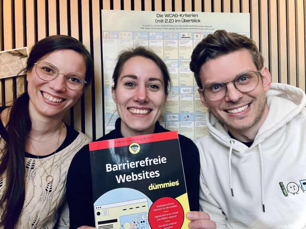 Zwei lachende junge Frauen mit langen braunen Haaren und ein junger Mann mit Brille und kurzen dunkelblonden Haaren, sie halten gemeinsam das Buch "Barrierefreie Websites für dummies" in der Hand