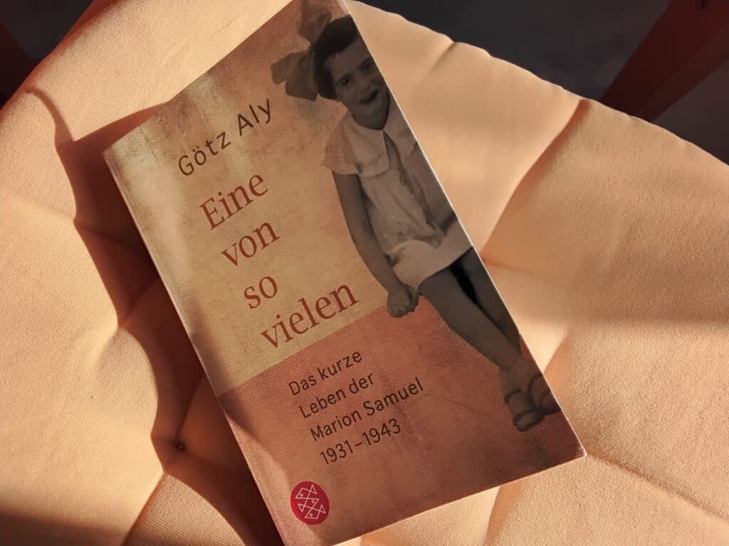 Buch "Eine von so vielen" auf einem orangefarbenen Kissen. Das Cover zeigt ein kleines Mädchen mit Pagenkopf, weißem Kleid und großer weißer Schleife auf dem Kopf.