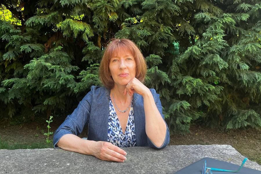 Ältere Frau mit einen rotbraunen Pagenkopf, blaugrauen Augen, einem blauweißen Kleid und einer Jeansjacke, sitzend an einem Gartentisch, dahinter Nadelgewächse.