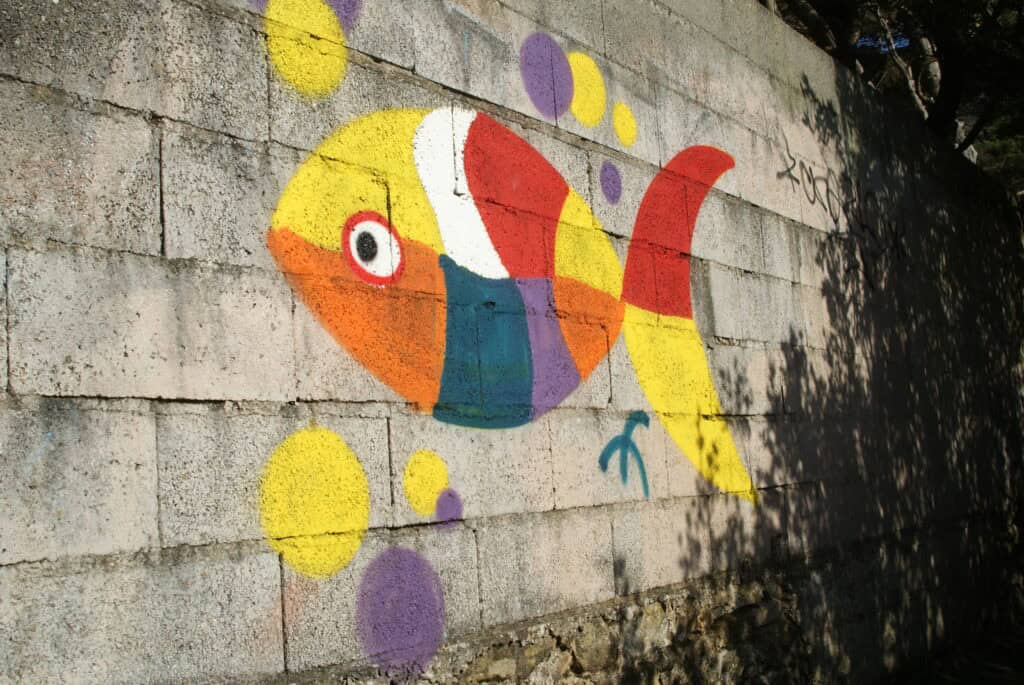 Fisch als Graffiti auf einer grauen Mauer. Der Fisch ist sehr bunt (gelb, weiß, rot, orange, grün, violett). Um den Fisch herum gelbe und violette Kreise.