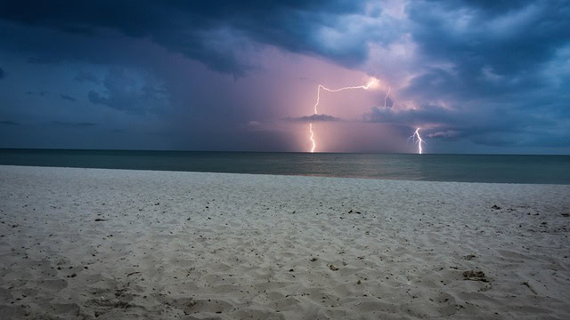 Im Vordergrund feiner Sandstrand, dahinter schwarzes Meer und Himmel mit schweren dunkelblauen Wolken, Blitze, um die Blitze herum ist der Himmel rosa